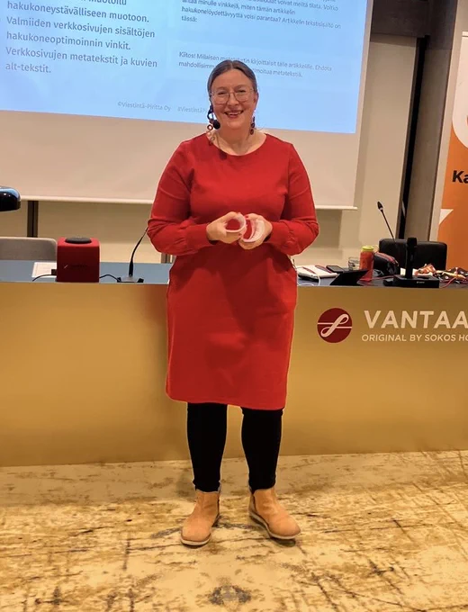 Nainen seisoo kokoustiloissa salin etuosassa ja hymyilee. Hänellä on yllään punainen mekko ja korvissa pitkät punaiset korvakorut. Päässään hänellä on headset-mikrofoni. Taustalla on pöytä, jonka etupaneelissa on teksti Original Sokos Hotel Vantaa. Pöydän taakse seinälle on heijastettu esitysdia, jossa on suomenkielistä tekstiä.