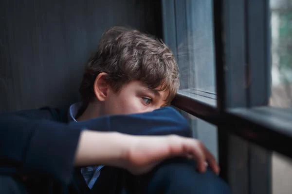 a teen boy sits and looks out a window with his face pressed into his arm