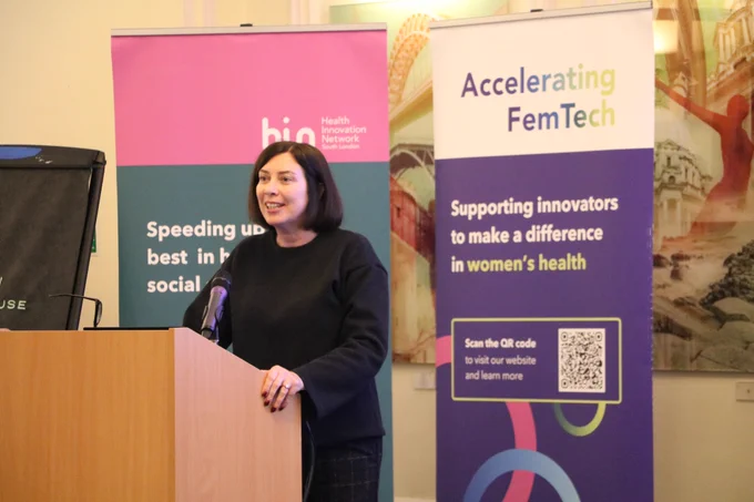 Speaker standing at a lectern with two banners behind, Health Innovation Network South London and Accelerating FemTech.