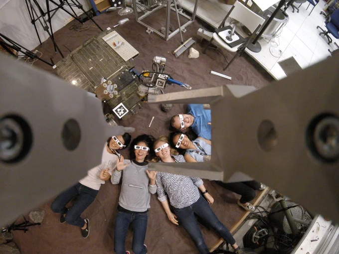 A group of NASA interns wear eclipse classes and lay on the ground looking upwards together. The group, in casual attire, all face the ceiling as a camera looks down at them positioned in a playful circle, behind them lab equipment covers the space. Credit: NASA