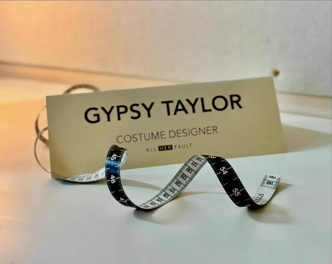 White background with a name card that says Gypsy Taylor: Costume Designer: All Her Fault. There is a measuring tape on the table as well.