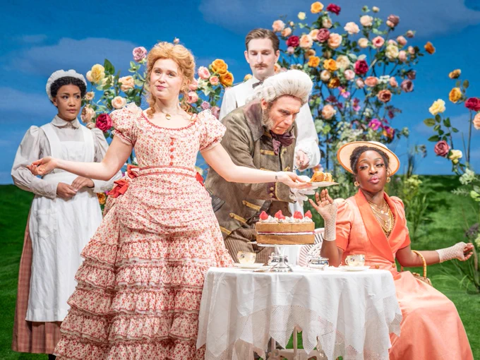 Five people in period costumes. One person is slicing a cake at a table set for tea, surrounded by an elaborate backdrop of colourful flowers.