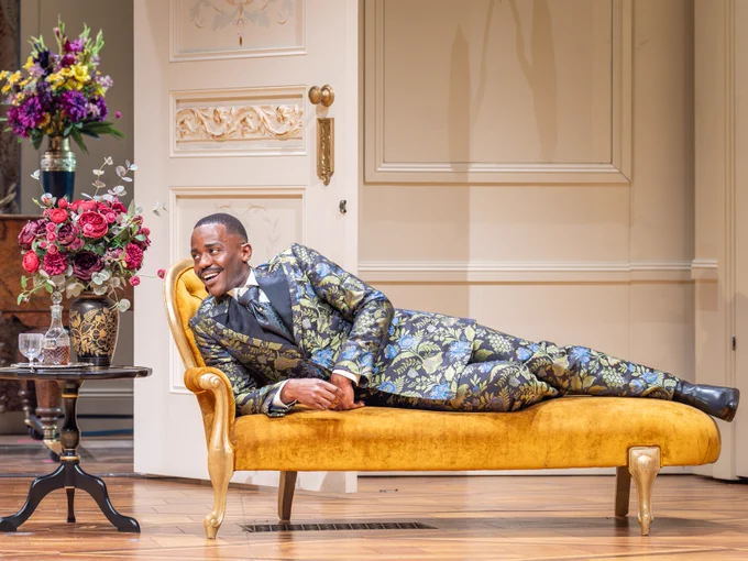 A man in a green, ornate floral suit reclines on a yellow chaise longue, smiling broadly, in a luxurious room with elegant decor. A vase of vibrant flowers is on a small table.
