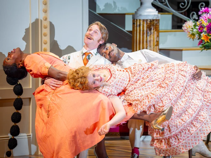 Four people sit on an elegant sofa. One person is holding another in a dramatic dip, while the other two excitingly watch the action.