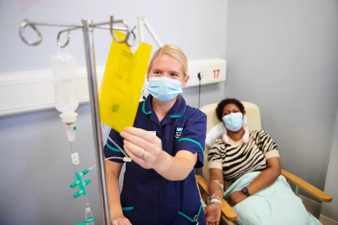 A healthcare worker in scrubs and a mask examining a medical document, with a patient sitting in the background, also masked, in a hospital setting.