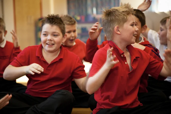 A group of children, mostly wearing red shirts, are sitting together and actively engaging in a cheerful activity. They are smiling, laughing, and gesturing animatedly at one another, showing a lively and interactive classroom atmosphere.