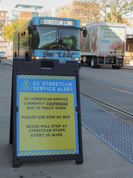 Kansas City Streetcar service alert sign indicating temporary changes due to track work, advising to use Ride KC bus with buses stopping every 20 minutes. In the background, a RideKC bus and a Sysco truck are visible on the street. It's a sunny day.