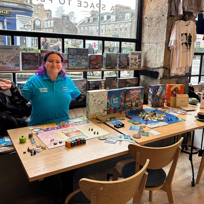 Game demonstrator from Asmodee in the Edinburgh board game shop Ancient Robot Games. They are smiling. In front of them is a table with the board games Inori, Fairy Ring and Survive the Island.