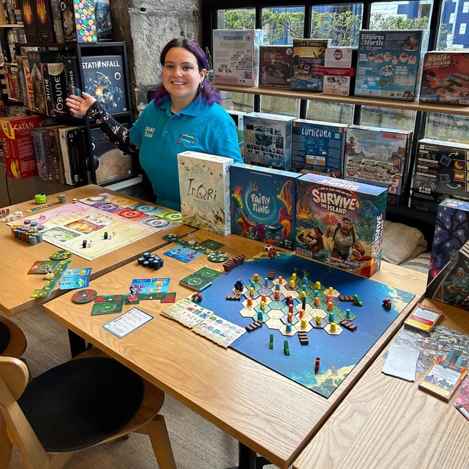 Demonstrator from Asmodee in the board game shop Ancient Robot Games. They are sitting behind a table with the board games Inori, Fairy Ring and Survive the Island.