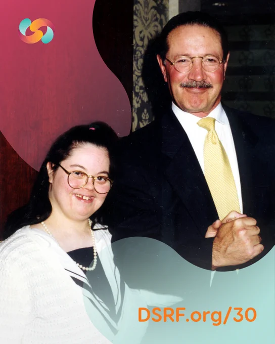 White woman with Down syndrome, who has dark hair half up and is wearing a formal white and black top with a pearl necklace, holds the hand of an older white man with a moustache and rounded glasses wearing a black suit with a yellow tie.
Text reads: DSRF.org/30