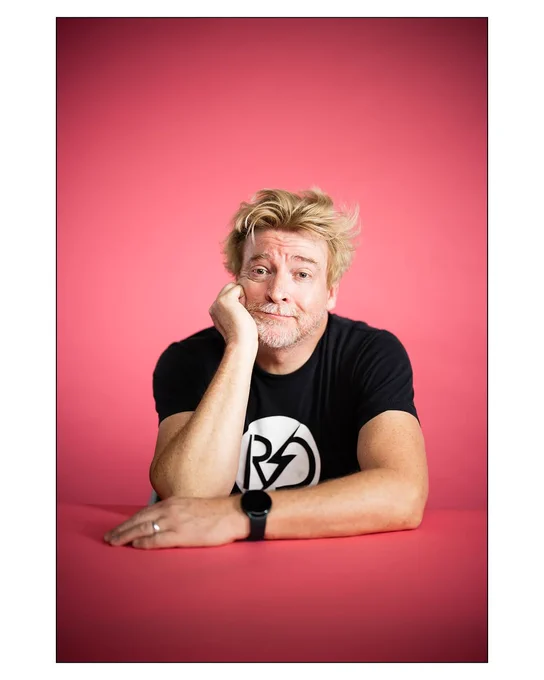 Rhys Darby in a black and white RD shirt leaning on a pink table with a pink background, one hand leans against his cheek and his golden hair is untamed.