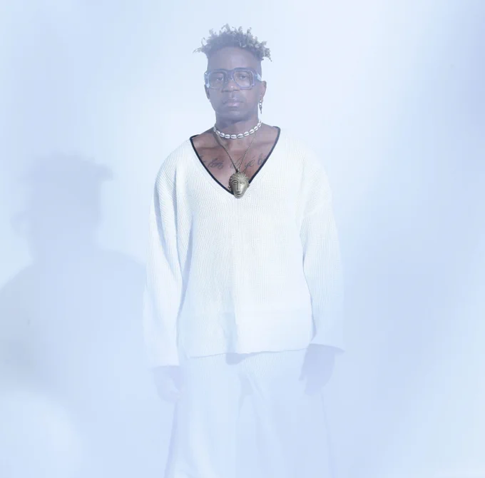 Cameroonian artist Blick Bassy stands looking at the camera wearing a loose fitted, long sleeved top, with a low v neck and long white trousers. He wears a tribal mask necklace in brass, a seashell choker necklace and blue thick framed glasses.
