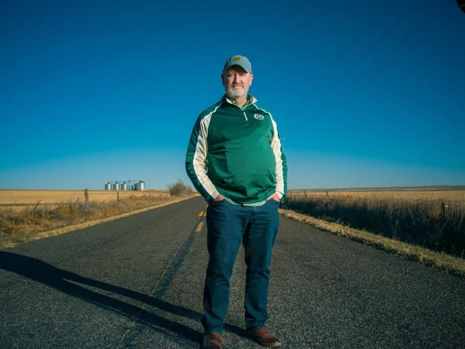 Jim Kuemmerle, a behavioral health specialist in eastern Colorado, poses for a photo with his hands in his pockets. He is wearing a green and gold Colorado State pullover, blue jeans, a faded green cap, and brown boots. He's standing in the middle of a road, while golden grasses, power lines, and a blue sky frame the background.