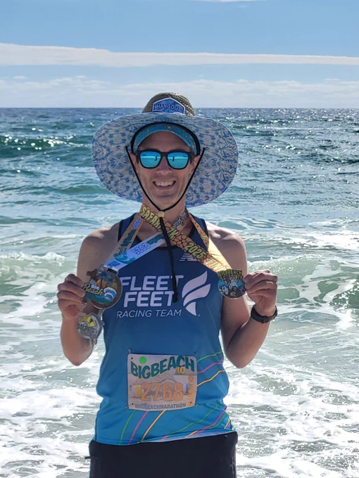 A white man wearing a turquoise running tank top that says, "Fleet Feet Racing Team," holds up multiple medals while standing on a beach with the surf washing up behind him.