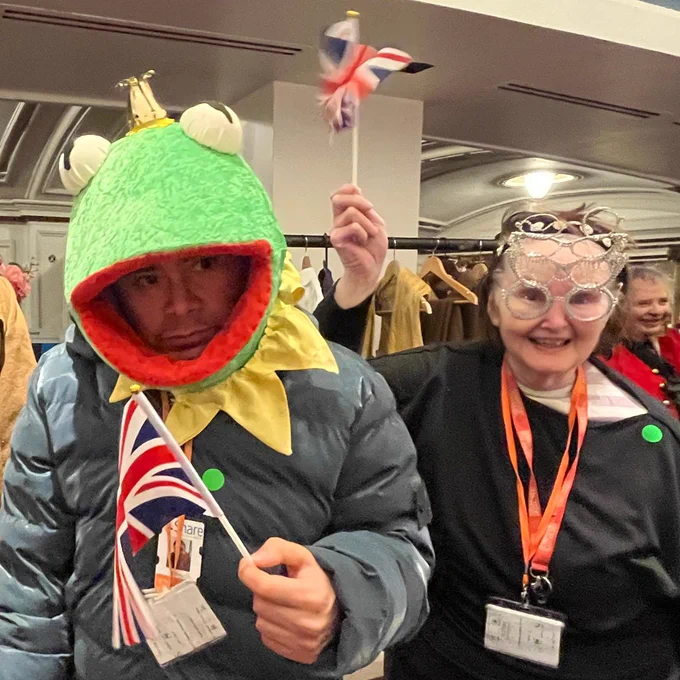 Two Share students dressed up in costumes and waving union jack flags. The student on the left is wearing a frog headpiece, and the student on the right is wearing masquerade-like silver glasses.