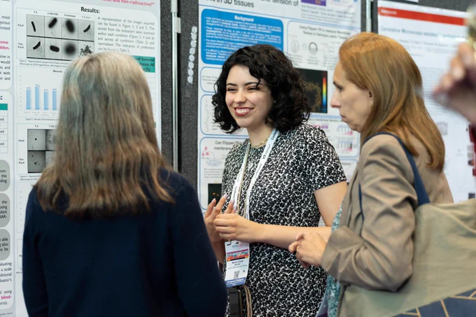 A delegate at mmc2023 smiles as she presents her scientific poster to two visitors during a poster session.