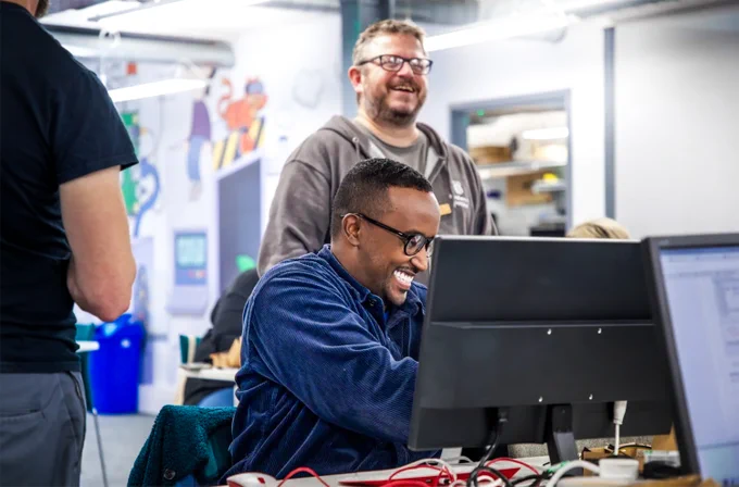 A man sat behind a computer screen laughing with a RPF trainer stood behind him. 