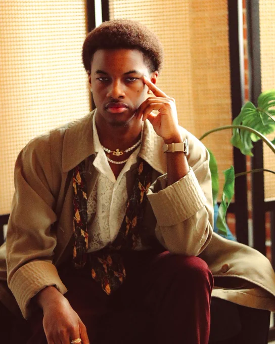 A stylish young person with short, natural hair sits in a relaxed pose, resting their hand on their face while gazing directly at the camera. They are wearing a layered outfit consisting of a lace shirt, a beige trench coat, and burgundy pants, accessorized with pearl necklaces and rings. The background features warm-toned woven textures and a green plant, creating a cozy and vintage-inspired atmosphere.