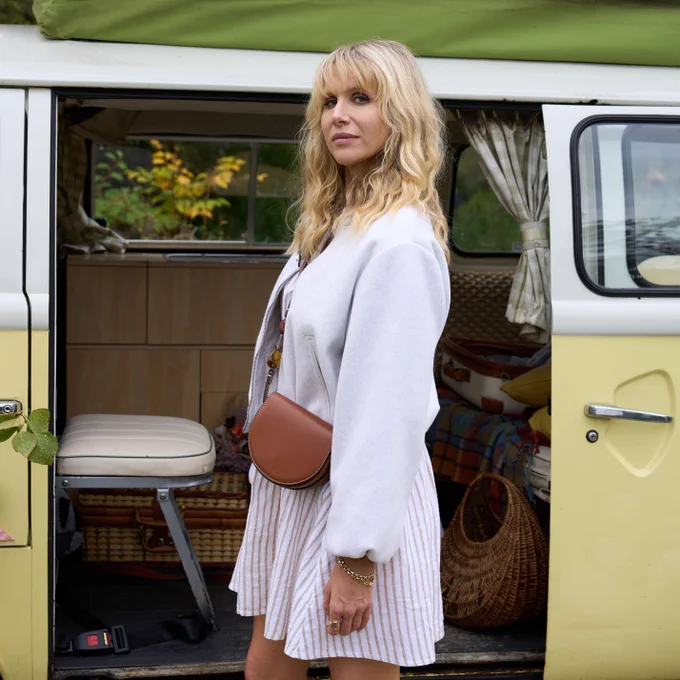 Amanda (Lucy Punch) stands in front of a yellow camper-van and looks to the camera. Wearing a white dress and jacket, she also has a cross-body brown leather handbag.