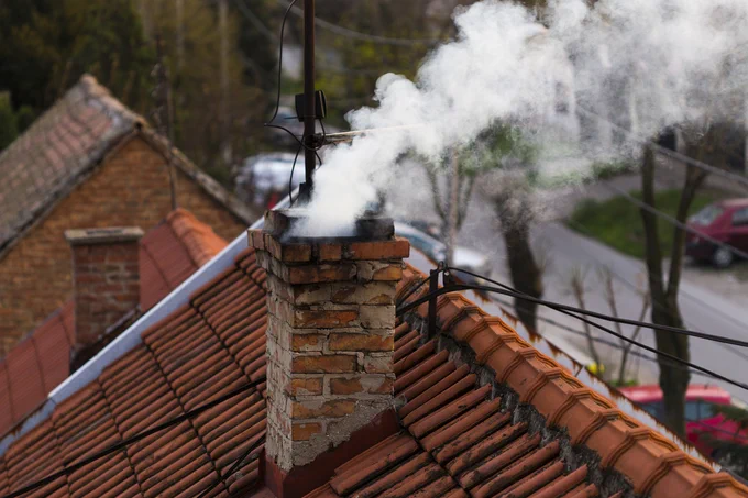A chimney on a tiled roof with white smoke coming out of it. The surrounding environment includes other houses and trees in the background.