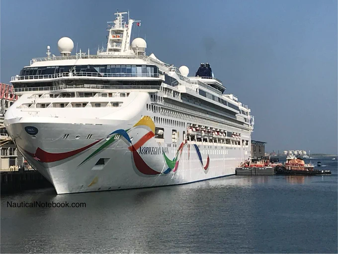 The cruise ship Norwegian Dawn docked at a cruise terminal, taken from a slightly angled frontal perspective, showing the bow and the starboard side of the cruise ship. The ship features a white hull with colorful wave- or ribbon like designs. A tug boat and floating platform are alongside. Photo credit Nautical notebook dot com.