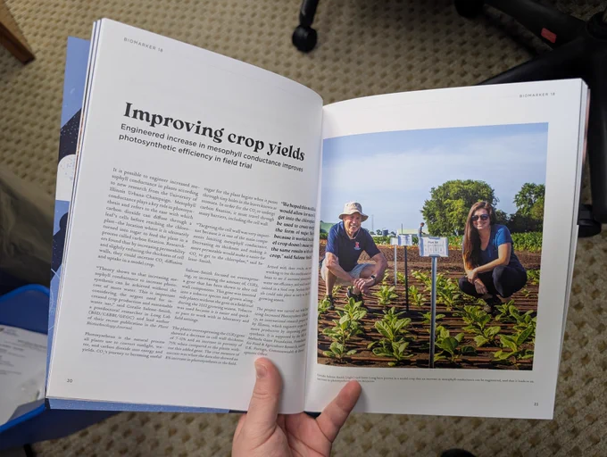 A magazine being held open. The left side has a headline that says Improving crop yields and a page of text. The right side of the magazine is a picture of two people kneeling in a field with young plants.