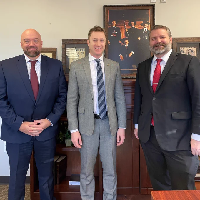 U.S. Attorney Josh Hurwit standing with First Assistant U.S. Attorney Justin Whatcott and Canyon County Prosecutor Chris Boyd. There are 4 framed photos in the background.