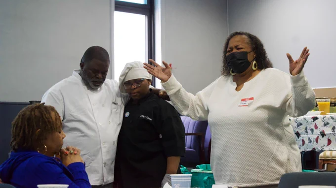 An older woman leads a prayer. Three people bow their heads in prayer.