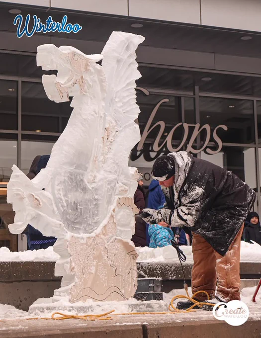 A person carving a large ice sculpture of a dragon at Winterloo outdoor event, surrounded by onlookers.