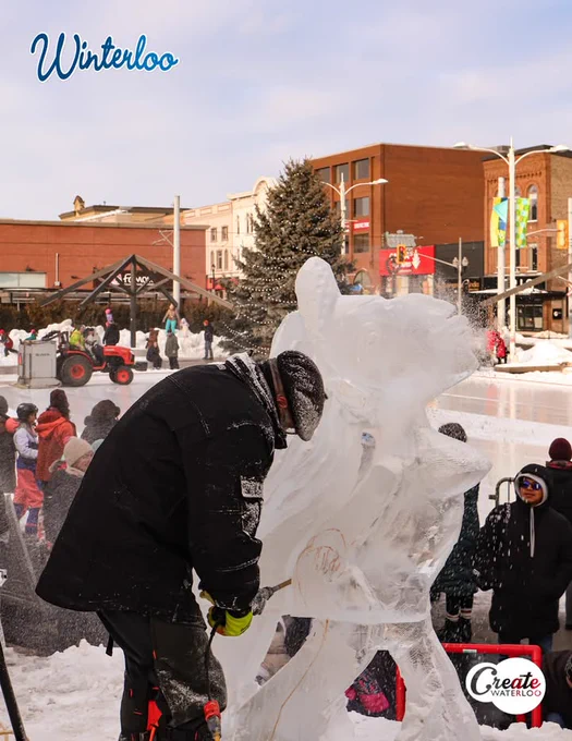 An artist carves an ice sculpture at the Winterloo Festival in Waterloo, with onlookers watching and an ice rink in background.