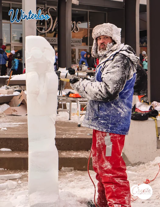 A person in winter clothing stands beside an unfinished ice sculpture at Winterloo event, surrounded by snow and equipped with sculpting tools.