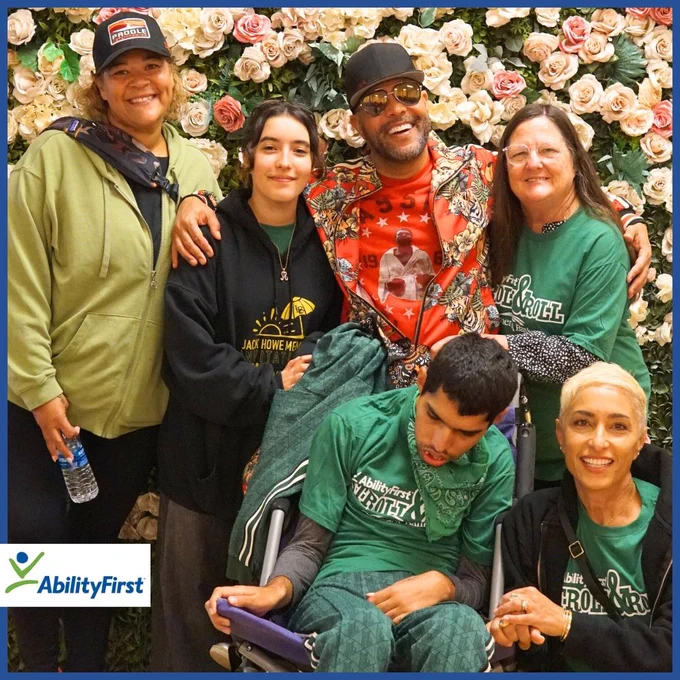 A diverse group of six smiling individuals, including one person in a wheelchair, posing in front of a vibrant flower wall. Several are wearing green shirts with 'AbilityFirst Stroll & Roll' printed on them, representing a joyful and inclusive event. The AbilityFirst logo is in the bottom-left corner.