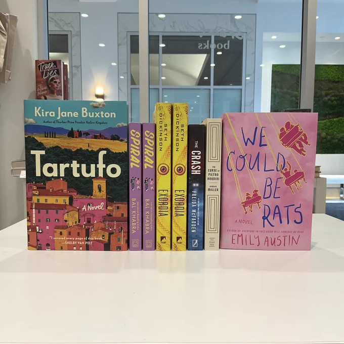 Horizontal book stack with titles such as “Tartufo” by Kira Jane Buxton; “Spiral” by Bal Khabra; “Exordia” by Beth Dickinson; “The Crash” by Freida McFadden; “The Curse of Pietro Houdini” by Derek B. Miller; and “We Could Be Rats” by Emily Austin.