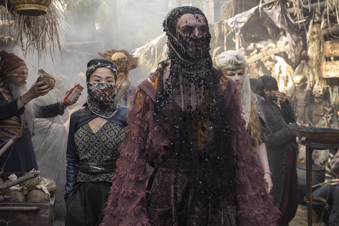 Mat Cauthon (Dónal Finn), Min Farshaw (Kae Alexander), and Elayne Trakand (Ceara Coveney) make their way through a bustling market in Tanchico. It is daytime, and all three are wearing veils.