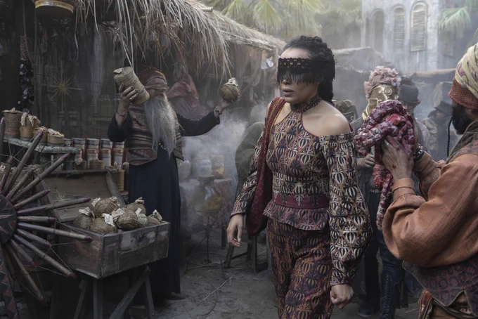 A veiled Nynaeve al'Meara (Zoë Robins) walks past vendors in a bustling Tanchico market during the day.