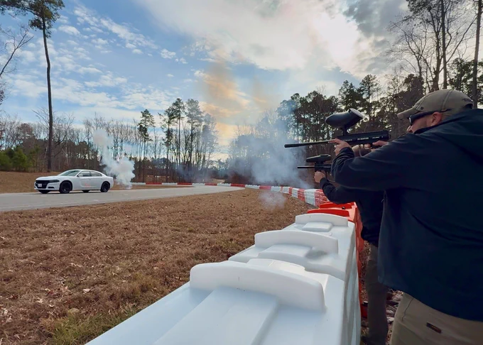 White car drives by loud explosives while being fired upon by structors.