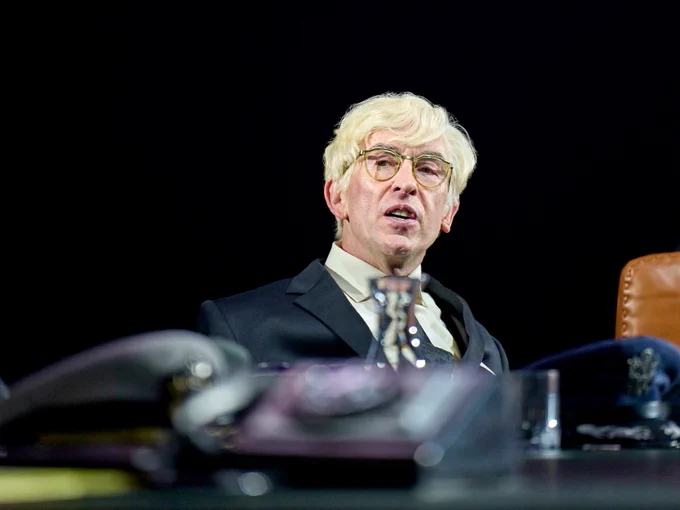 A man wearing a white wig and suit with a tie sits at a large desk.