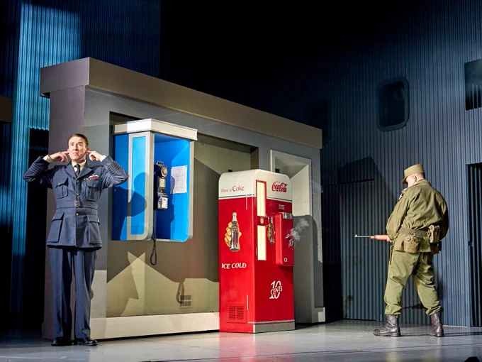 Two men in military uniforms stand on a stage with a phone booth and a vending machine in the background. One shoots towards the vending machine.