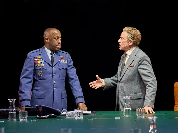 Two men stand on stage. One is dressed in a military uniform and the other in a business suit, engaged in a serious conversation at a table with glasses of water.