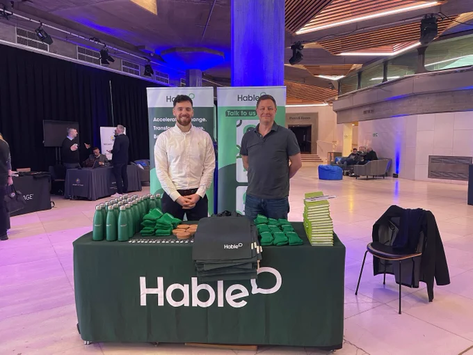 Two men stood behind a table in an industrial looking room with a green tablecloth on it that says "Hable." On top of the table is a number of different green products, including water bottles and tote bags.