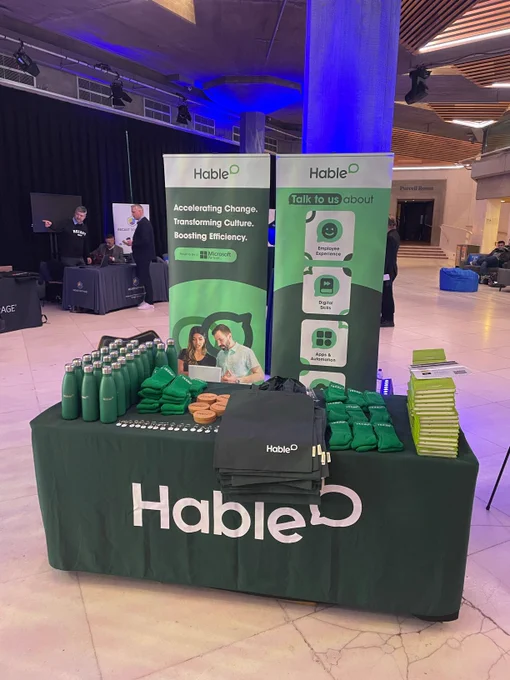 Table in an industrial looking room  with a green tablecloth on it that says "Hable." On top of the table is a number of different green products, including water bottles and tote bags.