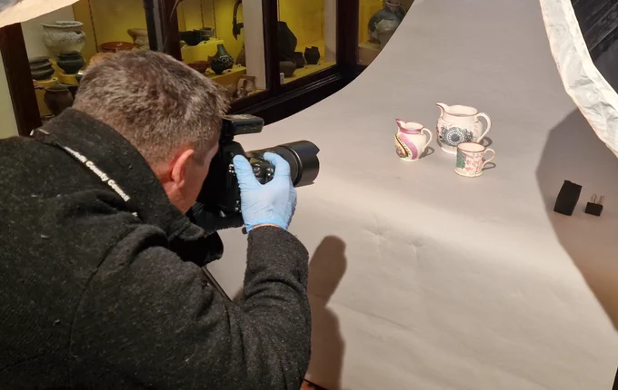Man holding a professional camera taking a photograph of three white, decorated pieces of china on a white background.