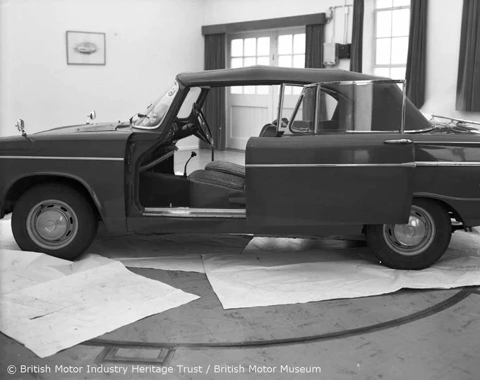 A black and white photograph showing the left side of a Hillman Super Minx with experimental sliding doors designed to open backwards. The vehicle is in a studio with both of its side doors open, showing some of the vehicle's interior. 