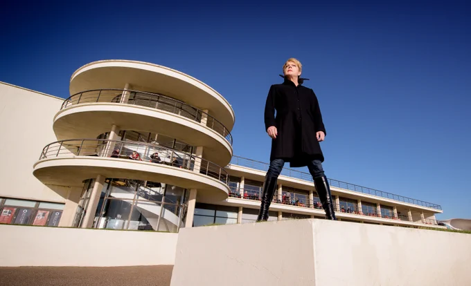 A person wearing black stands in front of a modernist building, the sky is blue and clear.