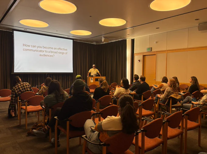 A seated audience attending a presentation titled "How can you become an effective communicator to a broad range of audiences?" Presenter Toby O'Geen stands at a podium next to the screen.