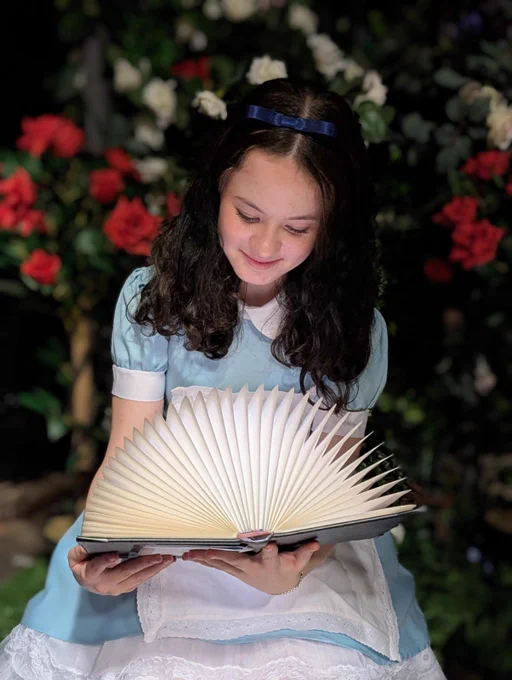 Student dressed in costume as Alice looking at a light up book, with red & white roses behind. She has long black hair and a blue hairband. 