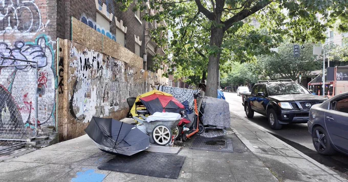 a street in PA.  A homeless person is living on the sidewalk, there is an open umbrella sitting upside down next to a tent an a cart. 