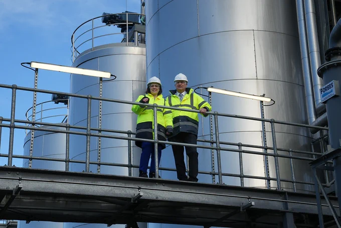 Image of the Deputy First Minister standing on a platform in a high visibility jacket, hard hat and protective glasses, next to the CEO of Celtic Renewables.