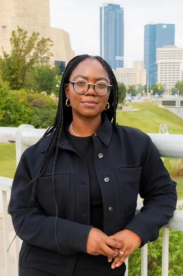 Photo of Chimwemwe standing in front of the Canadian Museum for Human Rights in Winnipeg. It is summer and she is standing on a footbridge. Her hair is in braids and go to mid-torso. She is wearing a black t shirt and a black button down jacket over top which is open. She has gold hoop earrings and round glasses that are a light yellow at the top and black on the bottom. She is slightly smiling. 