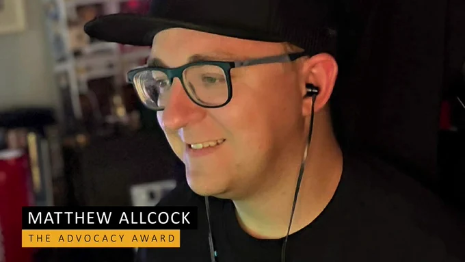 Matthew sat at his gaming desk wearing a cap, glasses and earphones, happily looking out of shot
